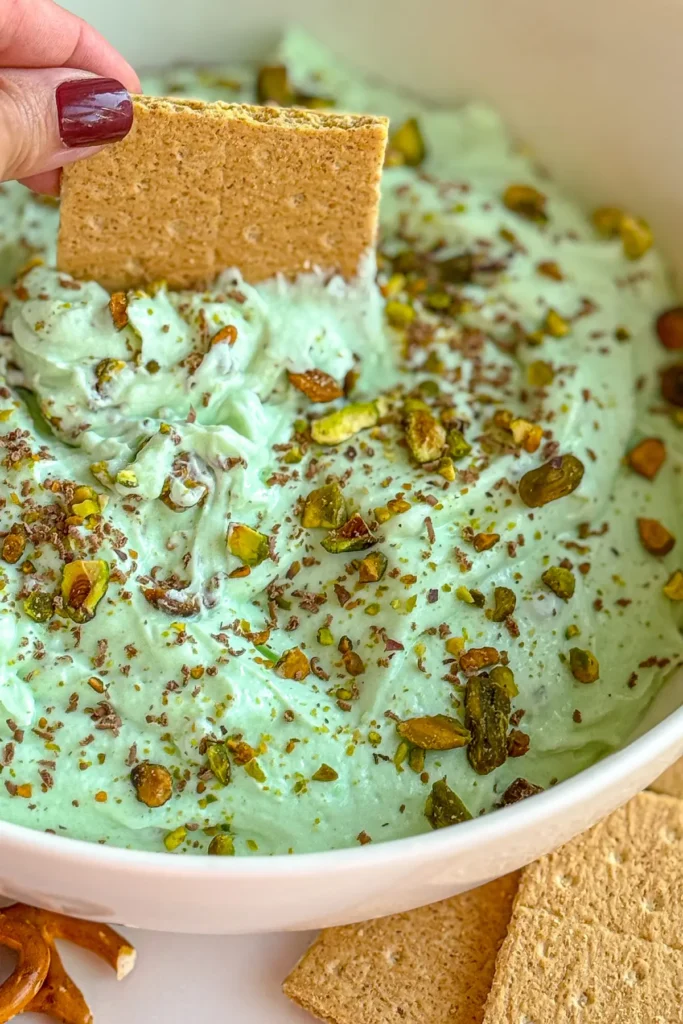 A close-up of pistachio pudding fluff topped with crushed pistachios and dark chocolate in a large white bowl and a graham cracker being dipped into it. 