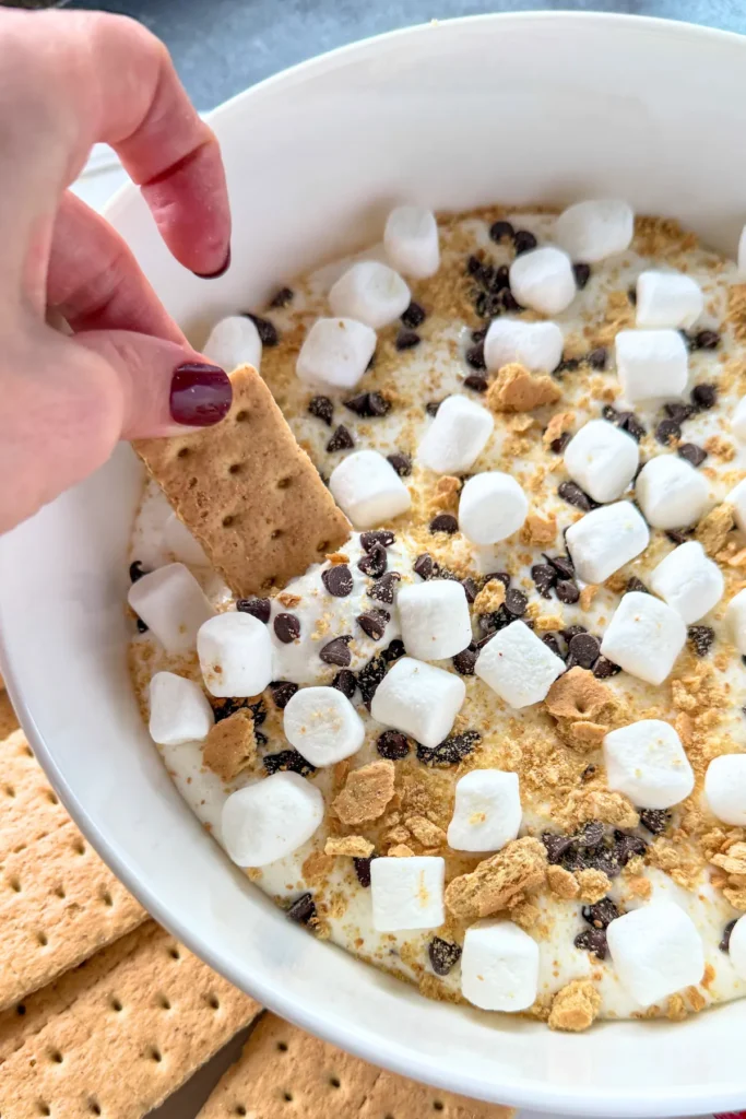 A graham cracker digging into cottage cheese s'mores dip topped with marshmallows and mini chocolate chips.