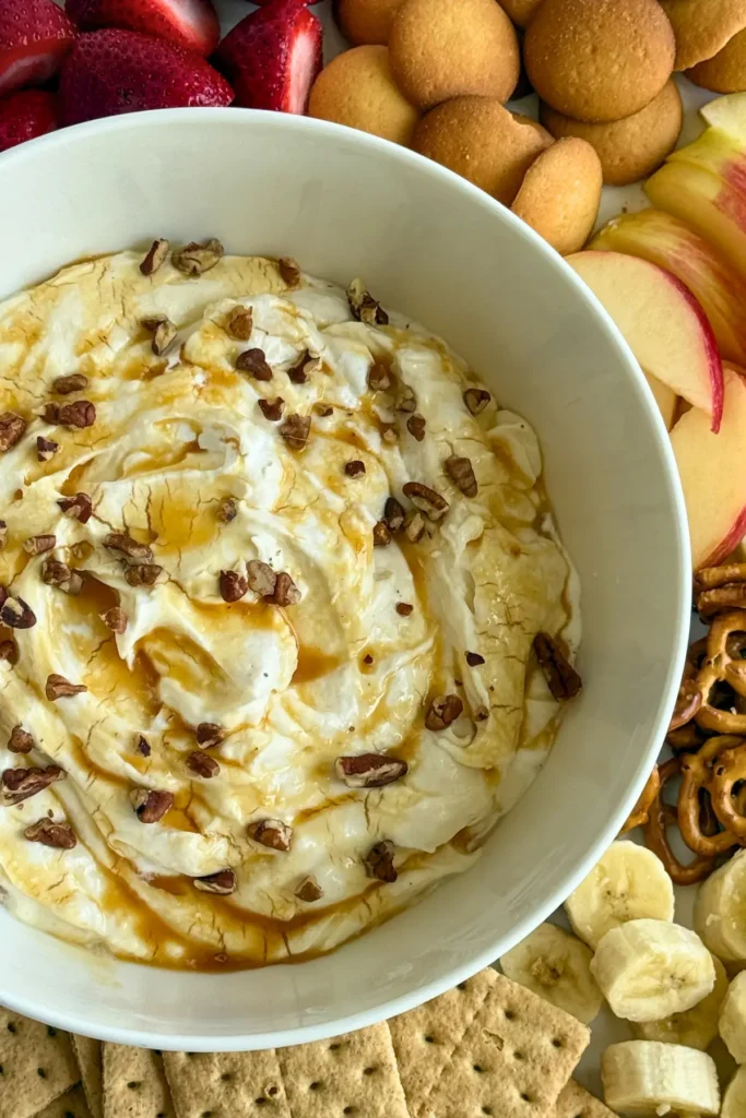 A large bowl of creamy Greek yogurt dip topped with pecans on a board with apple slices, strawberries, graham crackers and banana slices.