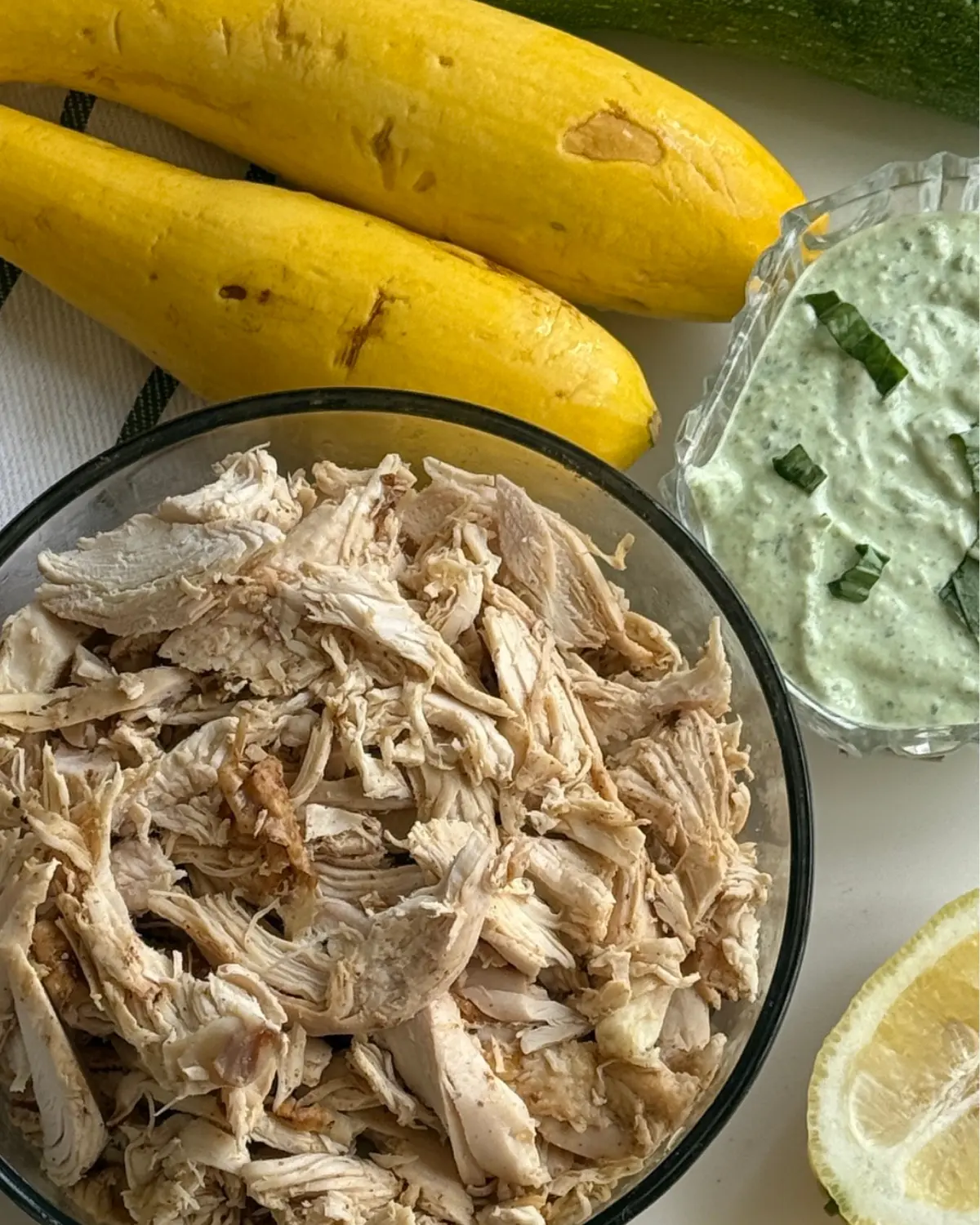 A bowl of shredded chicken next to lemon wedges and fresh summer squash on a counter.