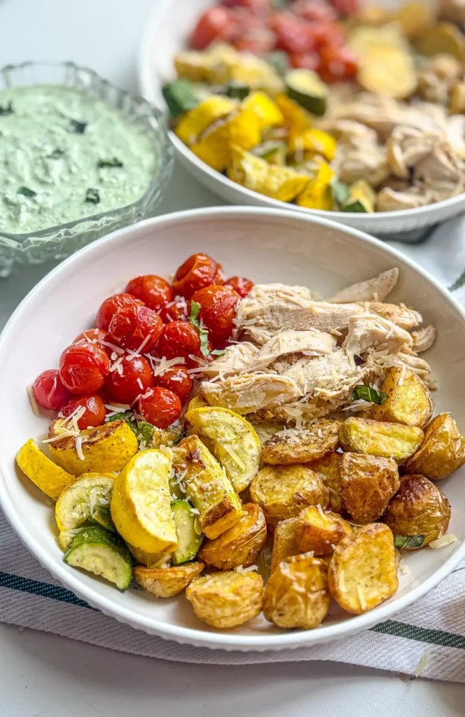 Roasted potatoes and veggies next to diced chicken in a bowl on a table next to the basil parmesan sauce.