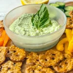 A bowl of parmesan basil Greek yogurt dip garnished with a basil leaf on a wooden cutting board surrounded by pretzels and bell pepper slices.