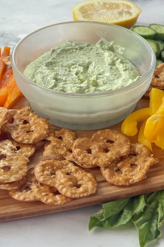 A bowl of parmesan basil dip on a board next to dippers like pretzels and bell peppers.
