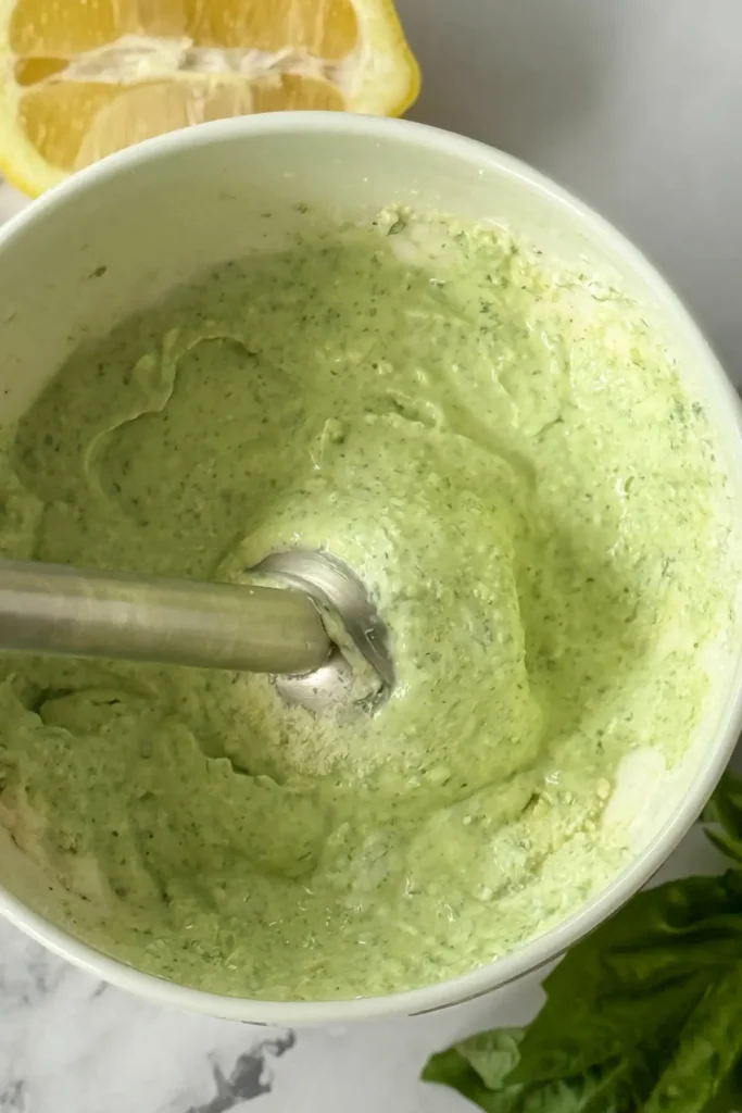 An immersion blender in a white bowl with ingredients for the basil parmesan dip pureed together.
