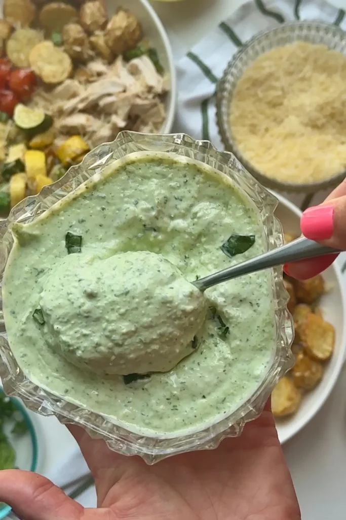Someone holding a bowl of parmesan basil Greek yogurt sauce holding a spoon of it in the other hands with parmesan roasted veggie bowls in the background.