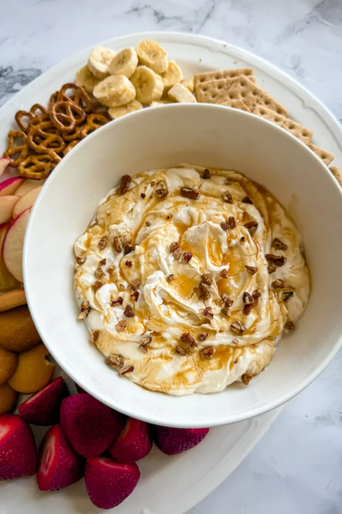 A birds-eye view of a large bowl of salted caramel cheesecake dip on a white platter surrounded by different dippers.