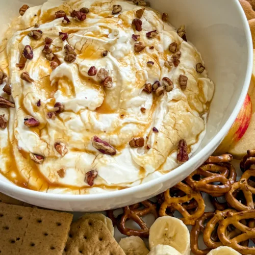 A large bowl of Greek yogurt salted cheesecake dip topped with chopped pecans surrounded by graham crackers, pretzels, banana slices, and strawberries.