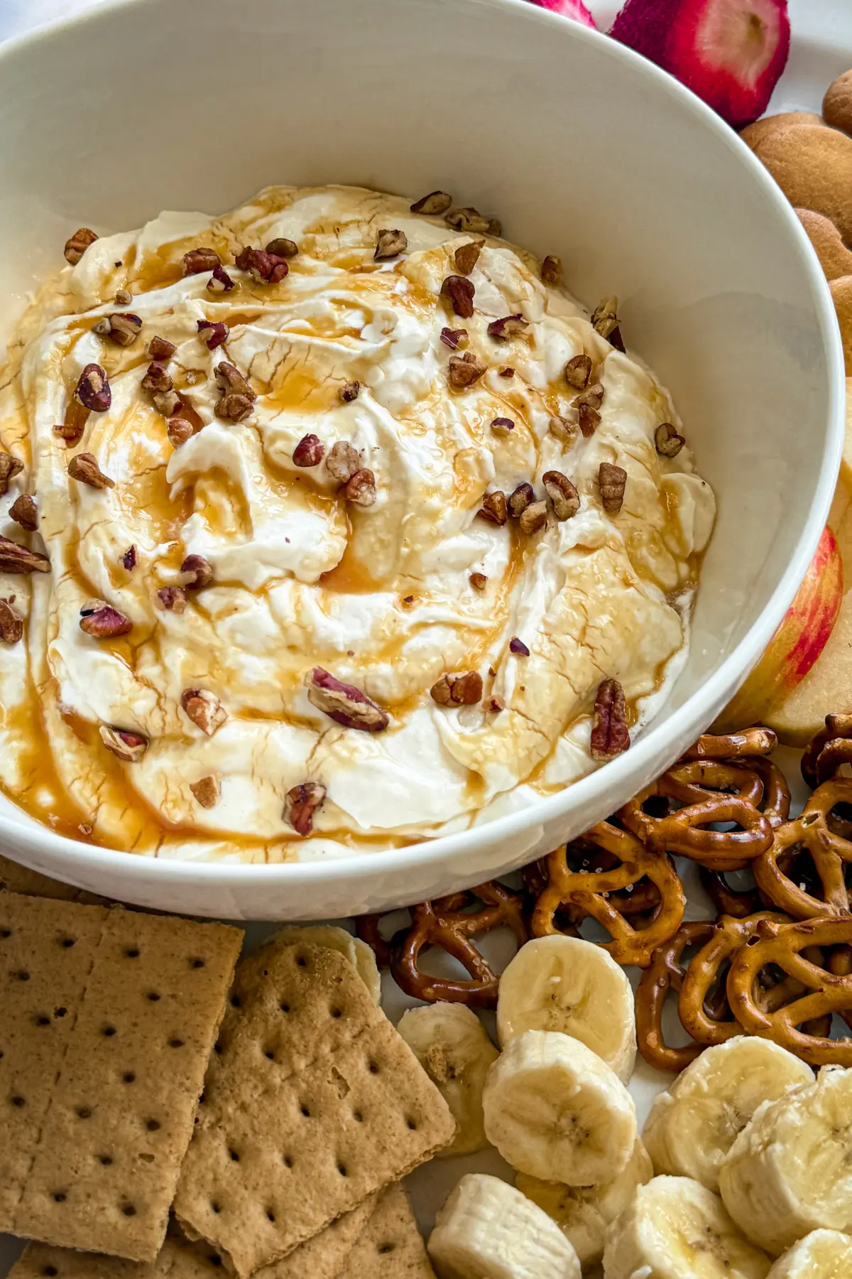 A large bowl of Greek yogurt salted cheesecake dip topped with chopped pecans surrounded by graham crackers, pretzels, banana slices, and strawberries.