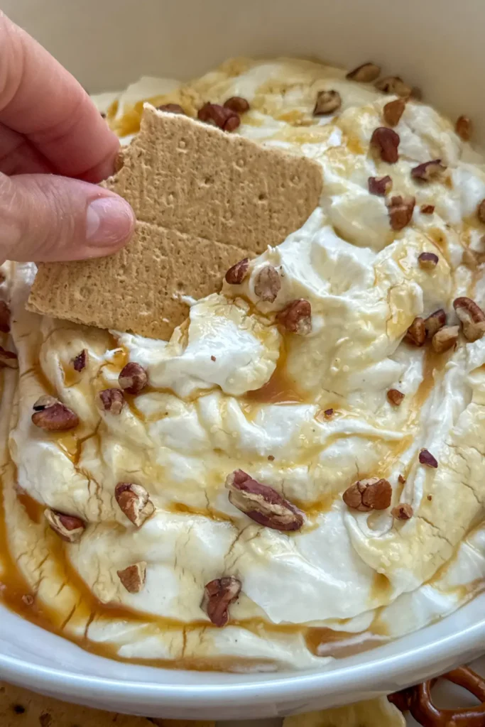 A graham cracker being dipped into a large bowl of caramel cheescake dip.