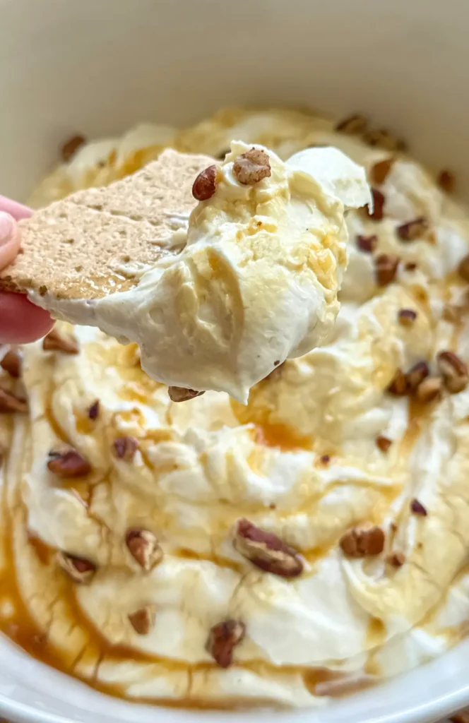 A graham cracker with fluffy salted caramel cheesecake dip over a big bowl of the dip.