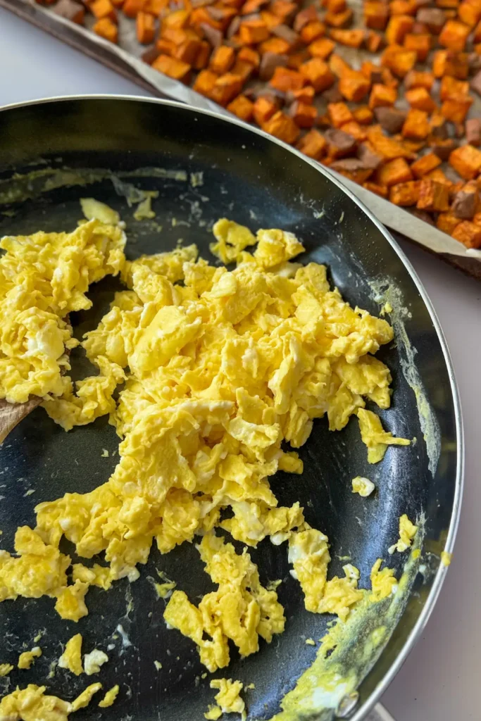 A pan of scrambled eggs next to a baking sheet with roasted sweet potatoes on it.