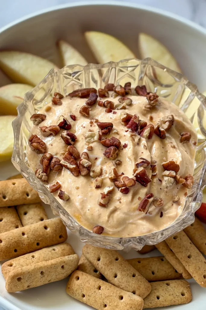A snack plate with a bowl of pumpkin yogurt topped with pecans, apple slices, and graham cracker sticks. 