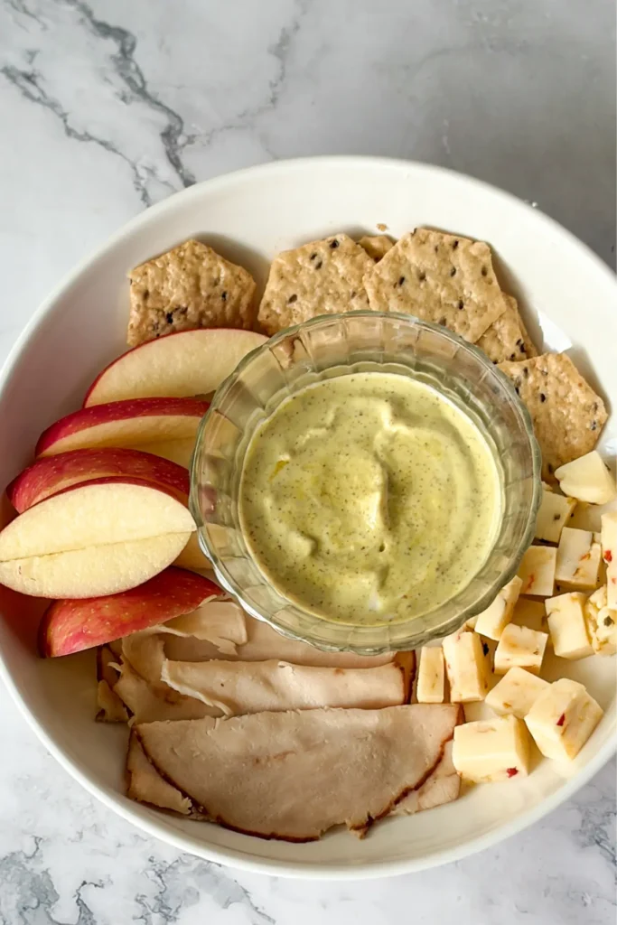 An apple, turkey, seeded cracker, and cheese plate with a bowl of maple Dijon yogurt dip in the middle. 