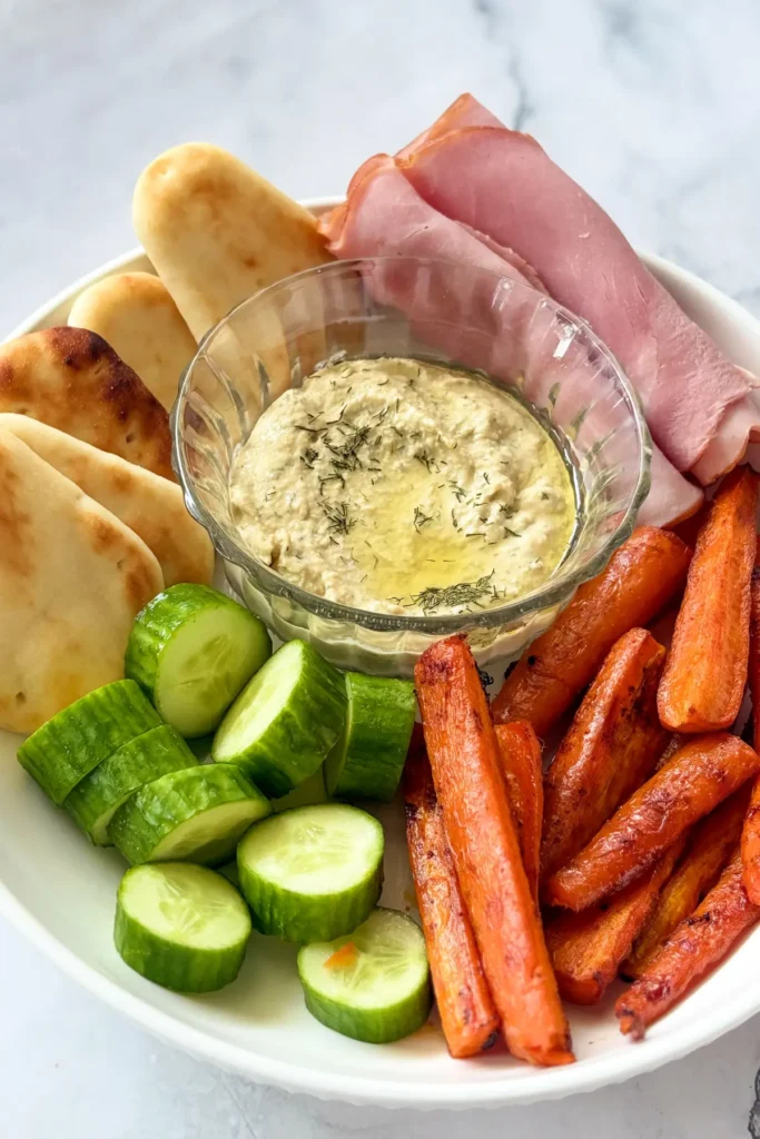 A snack plate with roasted carrots, diced cucumbers, naan dippers, ham, and a bowl of hummus.
