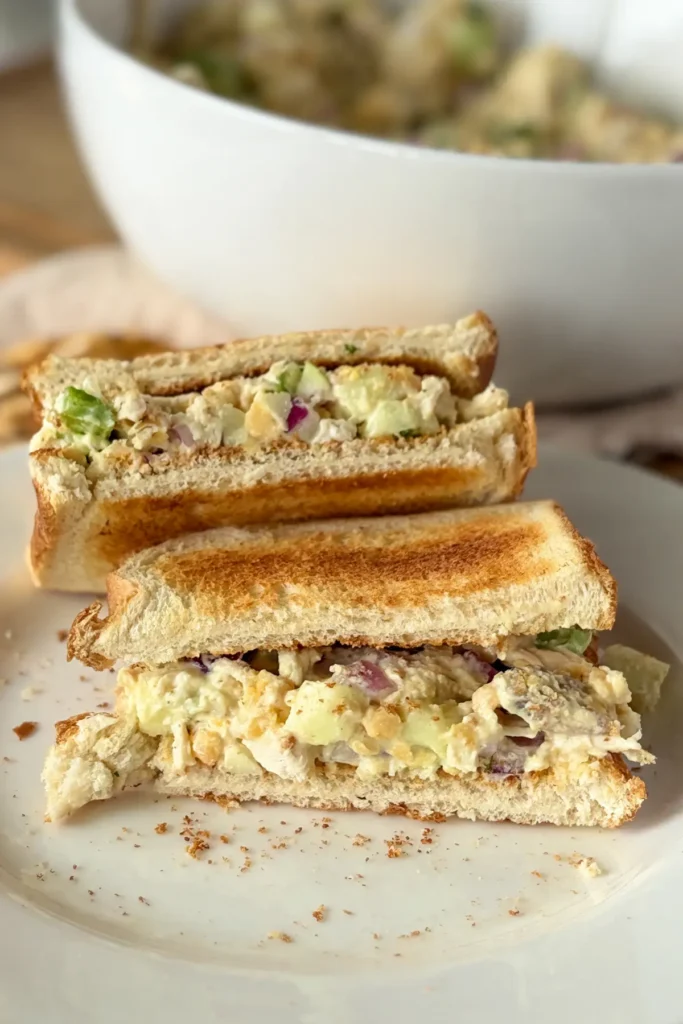 An image of a chicken salad sandwich showing the insides of the sandwich on a white plate next to a large bowl of chicken salad.