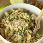 A bowl of chicken salad with a wooden spoon in it on a wooden cutting obard next to a half-eaten apple.