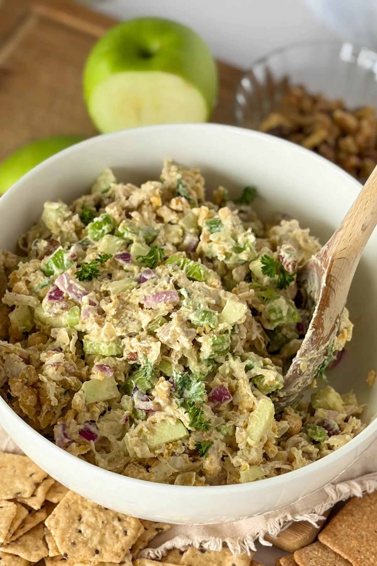 A bowl of chicken salad with a wooden spoon in it on a wooden cutting obard next to a half-eaten apple.