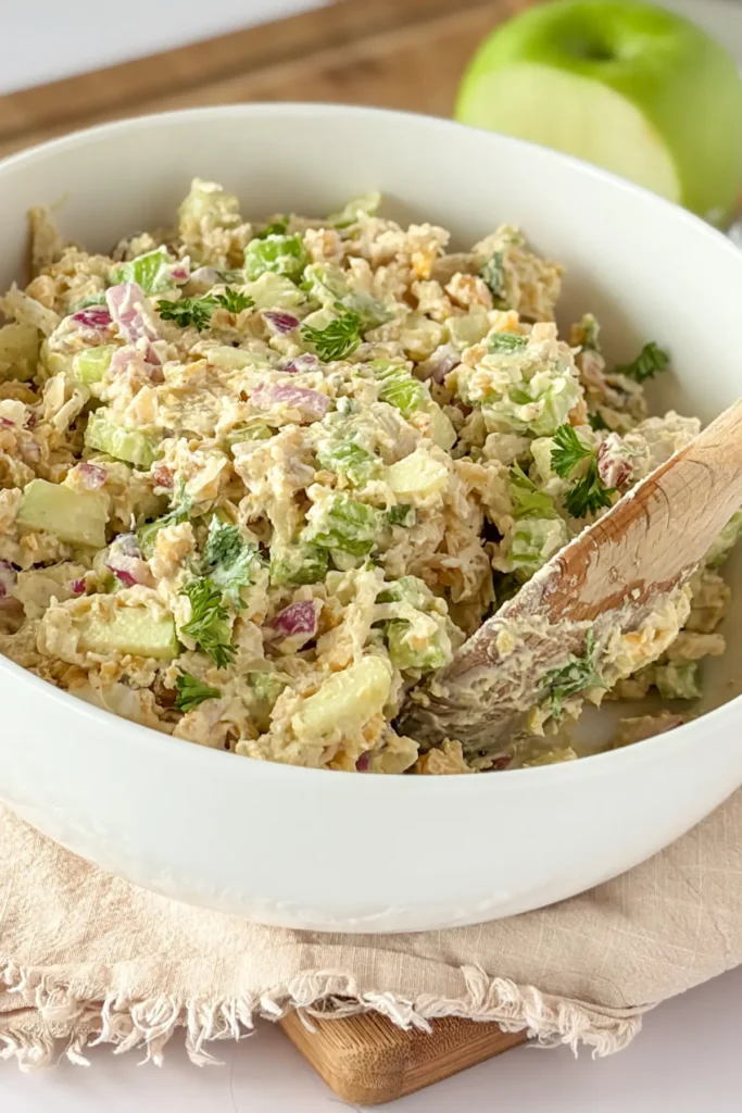 A bowl of chicken salad with a wooden spoon in it on a wooden cutting obard next to a half-eaten apple.