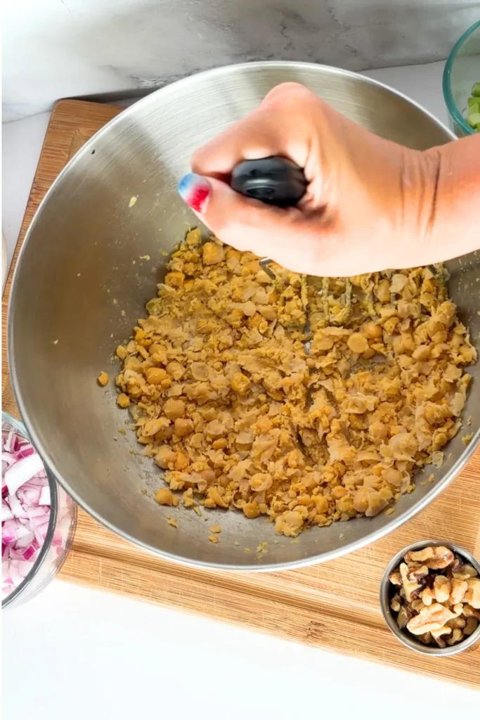 Someone using a potato masher to mash the chickpeas at the bottom of a mixing bowl on a counter.