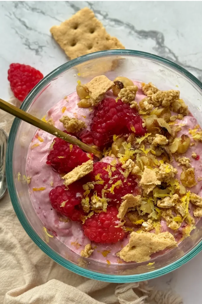 A bowl of lemon protein whip topped with chopped nuts, raspberries, graham crackers and lemon zest on a white counter.