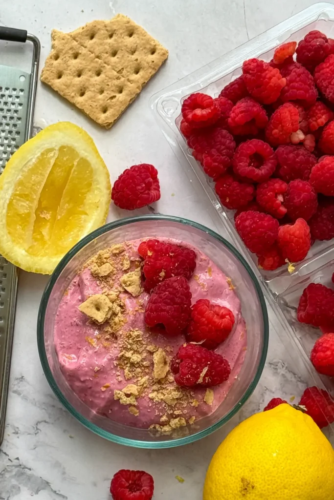 A birds-eye view of a bowl of raspberry protein whip topped with fresh raspberries and graham cracker crumbs on a counter next to a bowl of raspberries and lemon wedges.