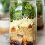 A maple balsamic chicken and grain salad jar on a counter with more salad jars in the background, without a lid on it.
