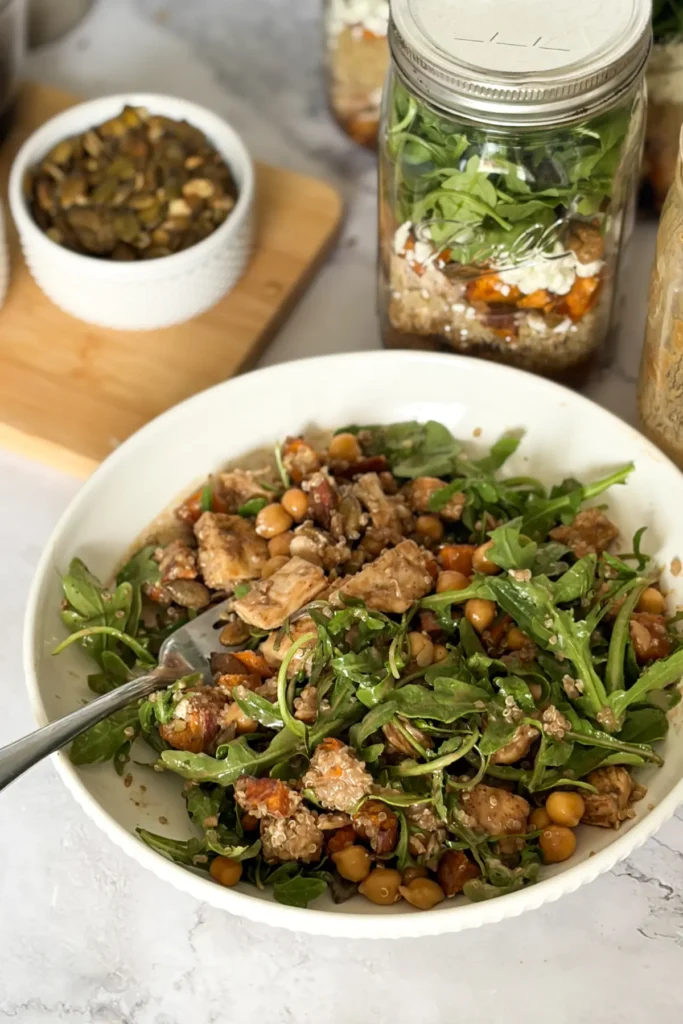 The maple balsamic chicken salad jar poured into a bowl with a fork next to a meal prep salad jar on a counter.