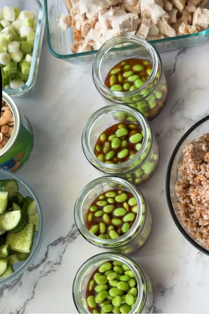 Four meal prep glass jars with sauce on the bottom and then edamame.