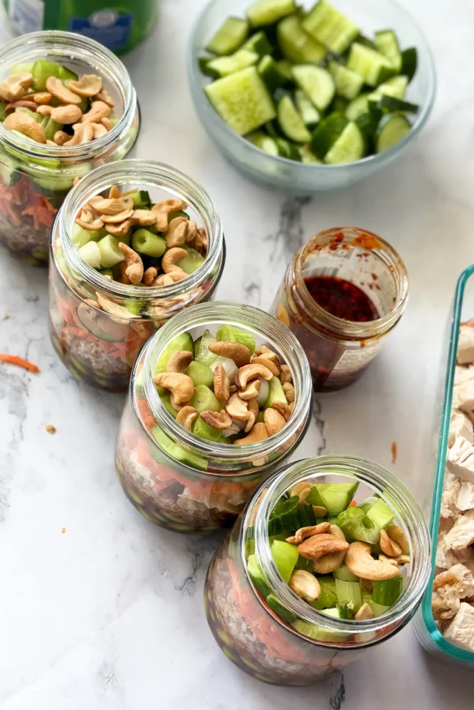 Four fully assembled meal prep salad jars lined up on a counter next to other salad jar components.