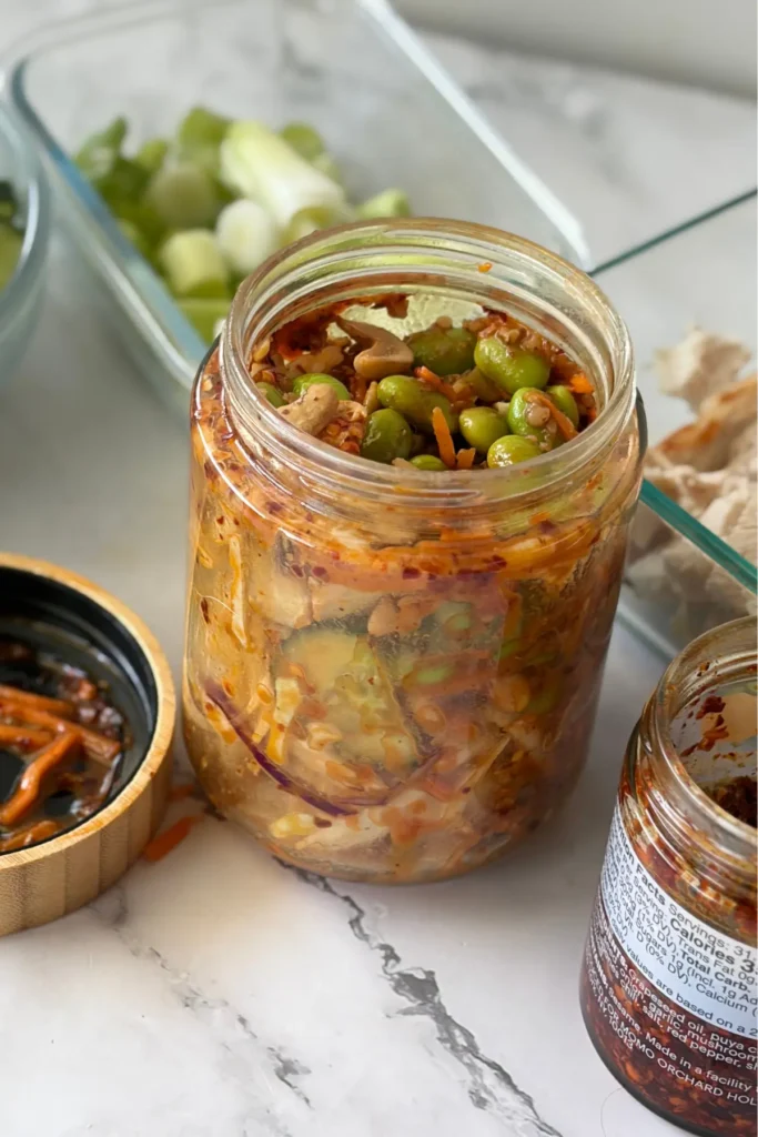 A shaken up salad jar on a counter next to chili crunch and cucumbers. 