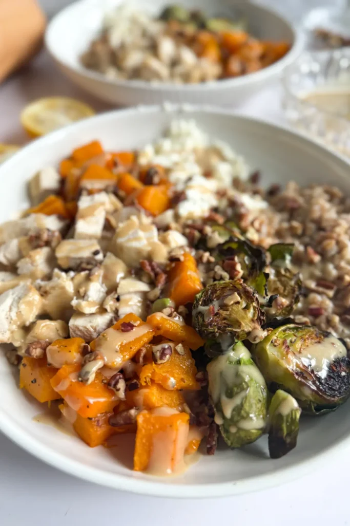 A fully assembled harvest bowl with chicken and drizzled with a maple tahini sauce on a table next to another plate.