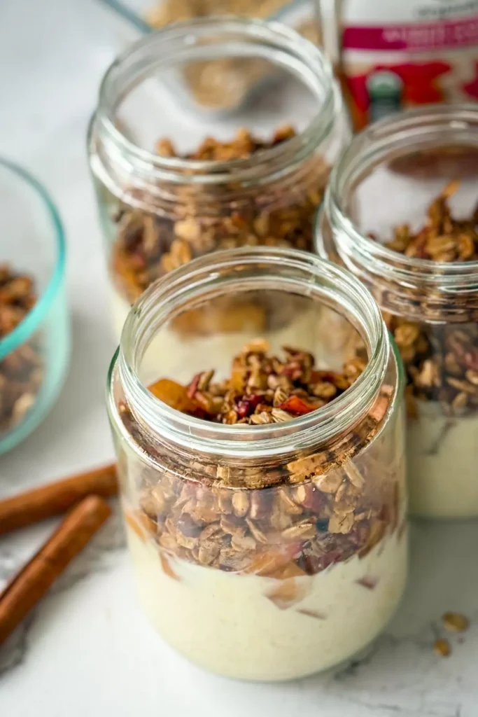 Three meal prep apple crisp breakfast jars assembled and on a counter next to cinnamon sticks.