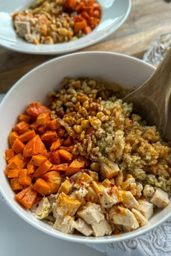 A large bowl showcasing the ingredients: diced sweet potatoes, quinoa, chicken, and nuts then drizzled with honey sriracha with a wooden spoon in it.