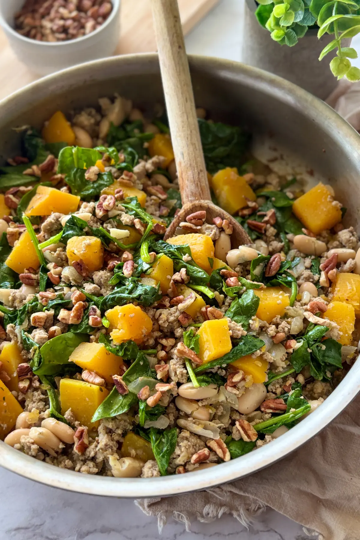 A large skillet with cooked ground turkey, butternut squash, spinach, and beans in it with a wooden spoon on a counter.