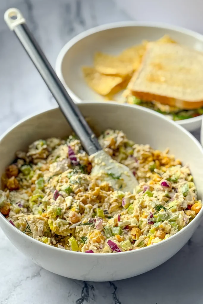 A large bowl of pepperoncini chicken salad with a spatula in it next to a plate with a chicken salad sandwich and chips on it.