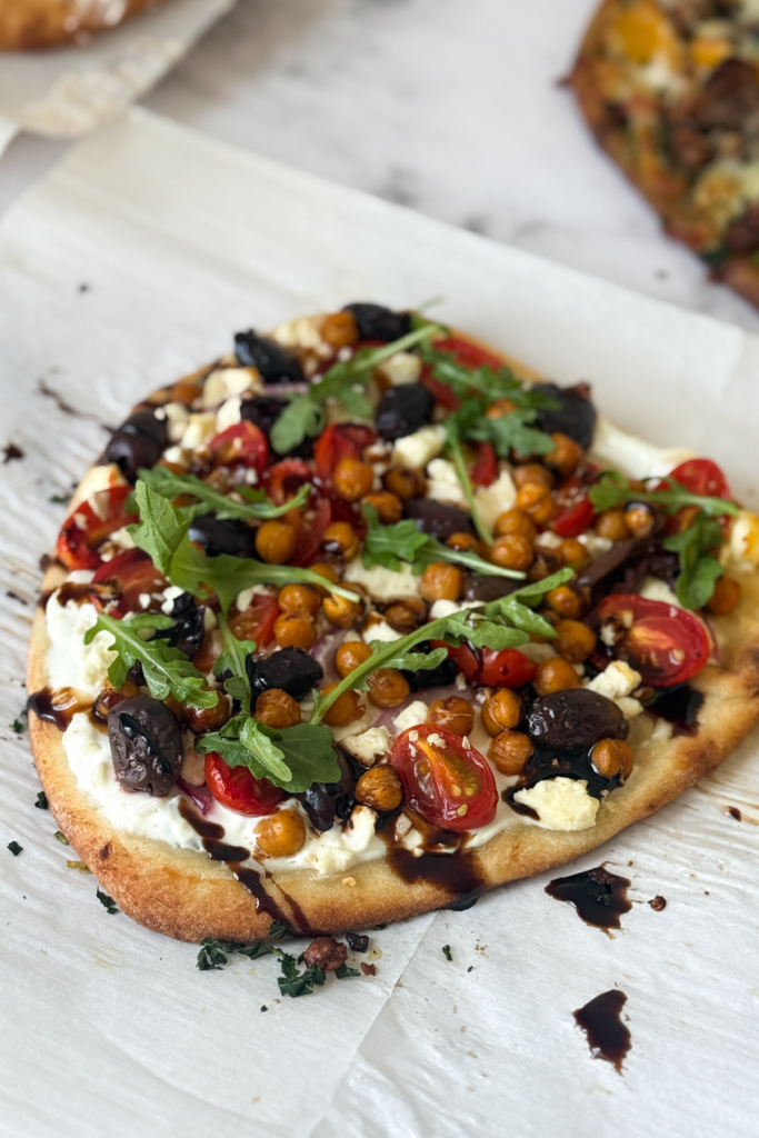 A Mediterranean chickpea and kalamata olive flatbread on parchment paper drizzled with balsamic glaze.