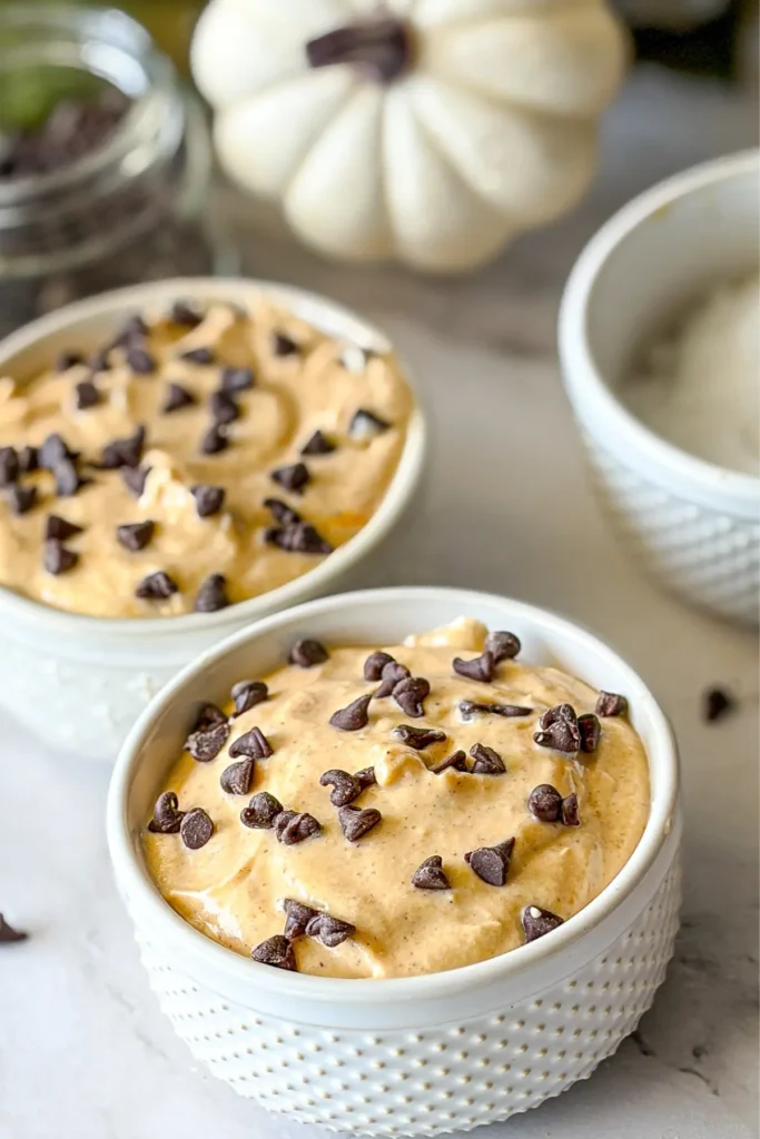 Two white ramekins full of the pumpkin cannoli dip with mini chocolate chips on top on a counter next to decorative pumpkins.