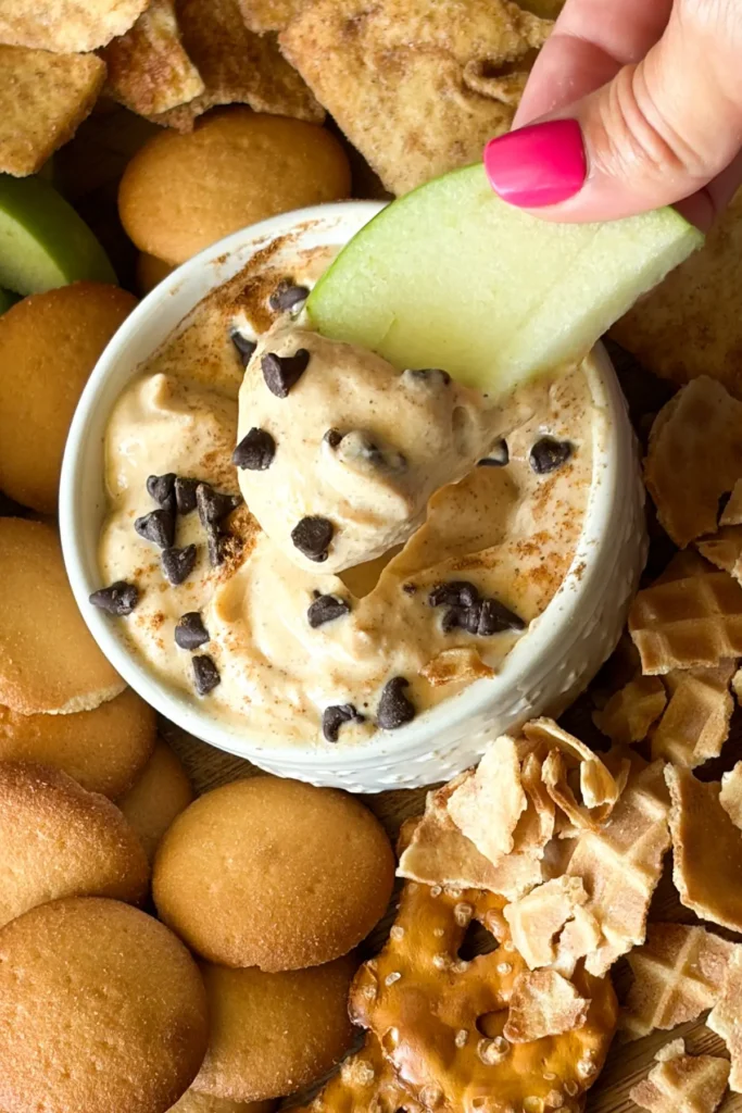 A Granny Smith apple slice dipped into the cannoli dip over a snack board.