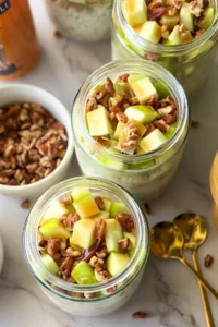 Meal prep jars with fully assembled salted caramel apple overnight oats and pecans on a counter next to spoons and a bowl of chopped pecans.