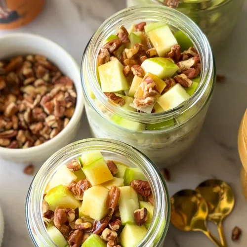 Meal prep jars with fully assembled salted caramel apple overnight oats and pecans on a counter next to spoons and a bowl of chopped pecans.