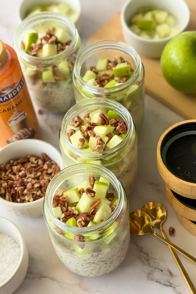 Four fully assembled caramel apple overnight oats with pecans and sea salt lined up on a counter next to the topping ingredients.