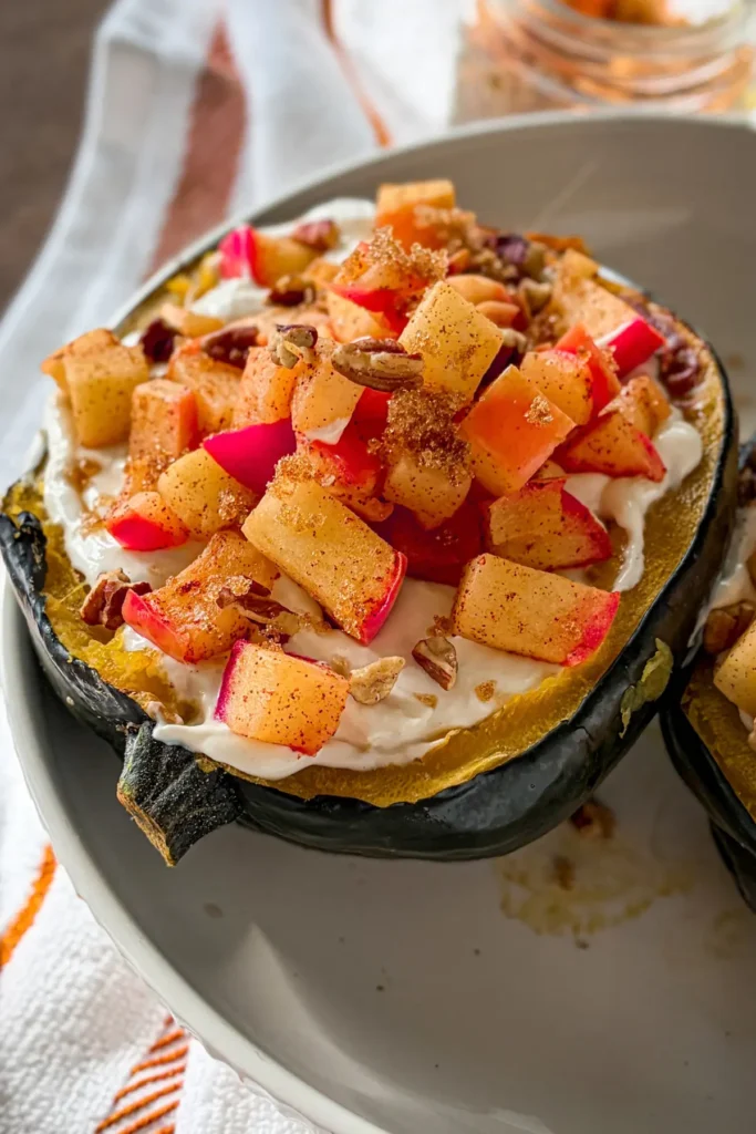 An acorn squash halved and topped with vanilla cheesecake filling, warm cinnamon apples. amd pecans on a white plate.
