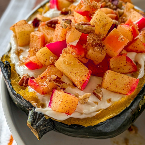 An acorn squash halved and topped with vanilla cheesecake filling, warm cinnamon apples. amd pecans on a white plate.