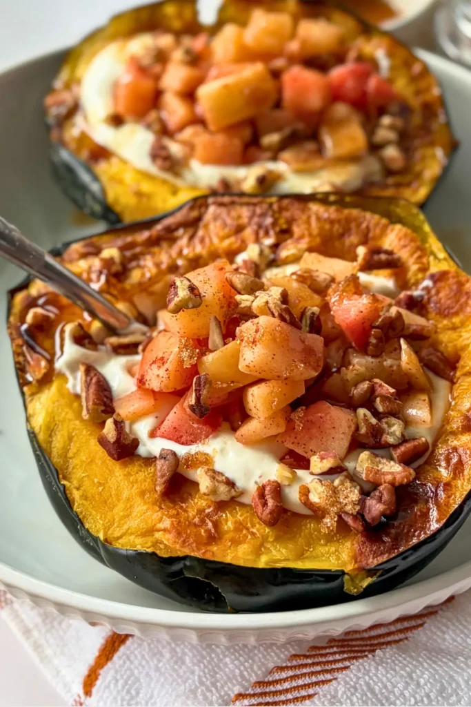 Two fully assembled cheesecake stuffed acorn squash topped with apples with a spoon in on on a plate.