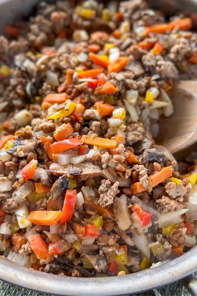 The ground beef, lentil, and veggie mixture cooking in a pan with a wooden spoon in it.