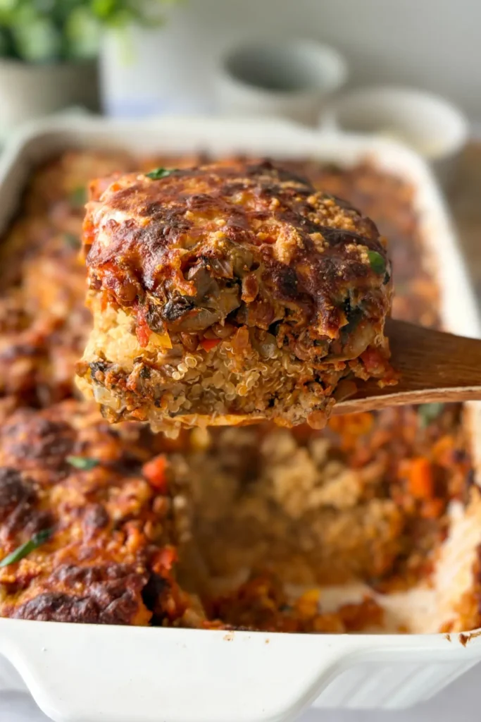 A large slice of the cheesy Italian quinoa casserole on a wooden spoon over a large glass casserole dish.