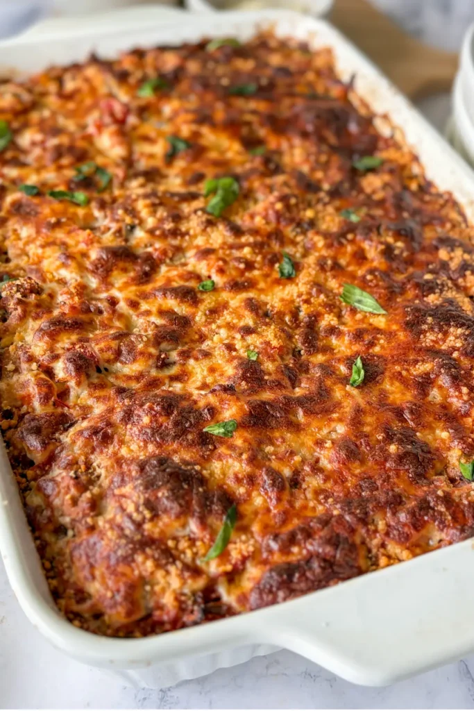 A crispy cheese Italian casserole in a white glass baking dish topped with basil leaves, right out of the oven, on a counter.