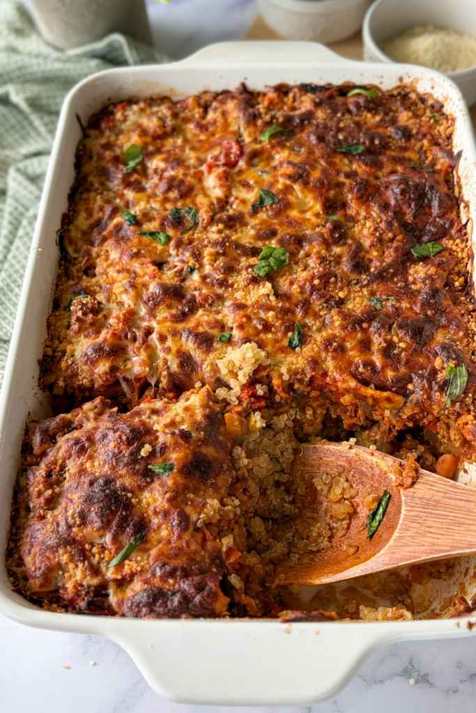 A wooden spoon digging into a crispy cheesy slice of casserole in the white glass baking dish on a counter.