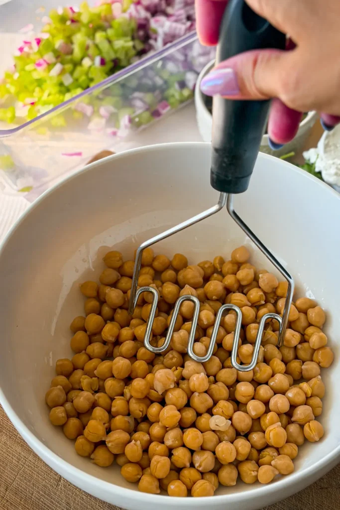 A potato masher mashing chickpeas in a large white bowl.