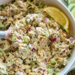 A birds-eye view of a large bowl of chicken piccata chicken salad garnished with lemon with a pink spatula in it on a counter..